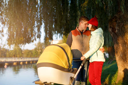 Young loving family walks by the lake with a stroller. Smiling parents couple with baby pram in autumn park. Love, parenthood, family, season and people concept.の写真素材