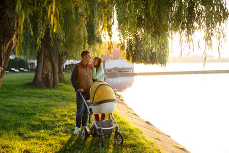 Young loving family walks by the lake with a stroller. Smiling parents couple with baby pram in...