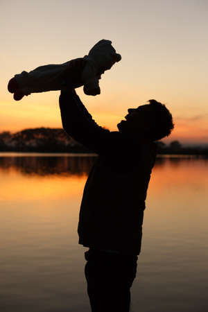 Silhouette of father holding little baby in his hands on sunset with lake on background. Man with child. Father's day theme.の写真素材