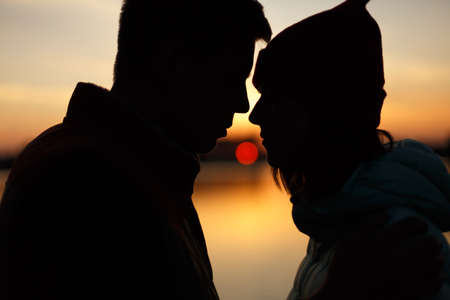 Silhouette of lovers couple on sunset by the lake. Sun between people profiles. Dusk sky, water on background. Passion in love concept, decoration, design for Valentines day.の写真素材