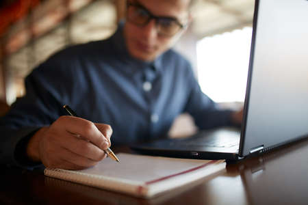 Close-up of man hand writing on paper notebook. Young businessman in glasses taking notes in notepad...
