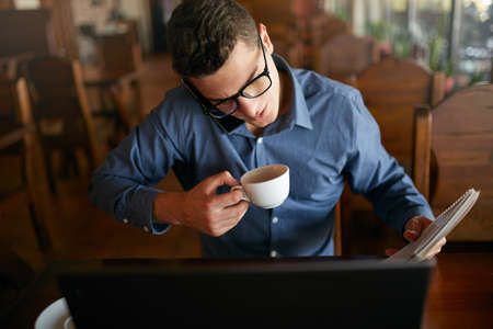 Overworked businessman speaking on cellphone holding it with shoulder, reading writings in notebook, drinking coffee and working on laptop simultaneously. Multitasking concept. Busy stylish man.の写真素材