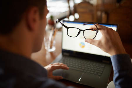 Back view of man hand holding eyeglasses in front of laptop screen with charts and diagrams. Poor eyesight threatment theme. Computer glasses. Break for eye rest.の写真素材