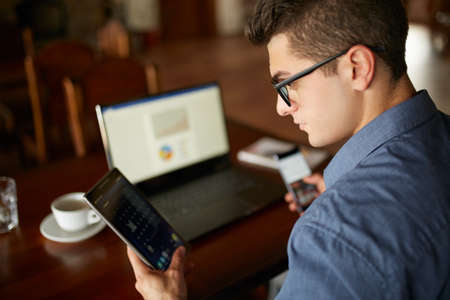 Attractive man in glasses working with multiple electronic internet devices. Freelancer businessman has laptop and smartphone in hands and laptop on table with charts on screen. Multitasking theme.の写真素材