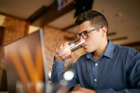 Handsome caucasian man with glass of water in hand works with laptop. Businessman in glasses drinks water for body hydration while working. Attractive designer quench thirst. Healthy lifestyle theme.の写真素材