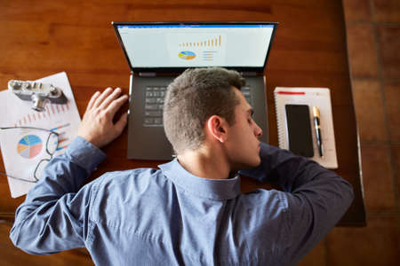 Top view of tired exhausted businessman sleeping on laptop keyboard at workplace. Handsome overworked freelancer man lying on wooden table and relaxes dreaming near notebook and documents with charts.の写真素材