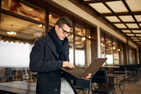 Handsome attractive businessman using a laptop on office terrace. Stylish man in glasses coat and scarf standing on balcony with laptop. Smart casual clothes. Business theme.の写真素材