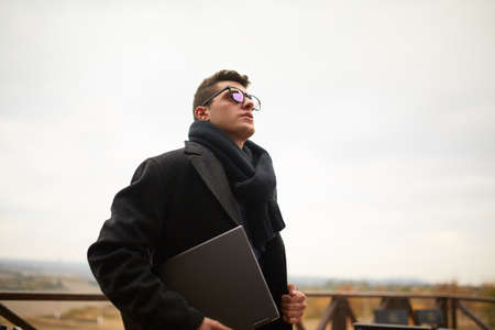 Handsome attractive businessman using a laptop on office terrace. Stylish man in glasses coat and scarf standing on balcony with laptop. Smart casual clothes. Business theme.の写真素材