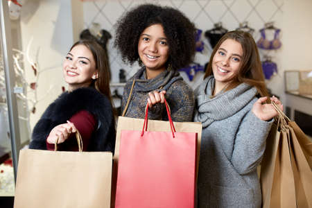 Women of diverse ethnicity with shopping bags posing in lingerie store. Portrait of three pretty multiracial girls walking in underwear shop on sale. Copyspace on large paper craft bags.の写真素材