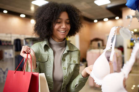 Young smiling attractive african american woman choosing right bra size in lingerie store. Black mixed race pretty girl choosing underwear in shop and holding paper shopping bags.の写真素材
