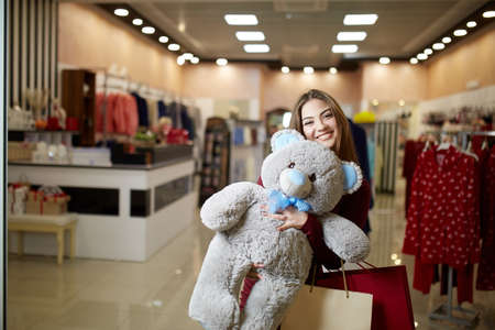 Attractive young cute caucasian woman hugs plush teddy bear with shopping bags at clothing store front. Pretty girl shopper happy with her purchases and prize in shop sale competition.の写真素材