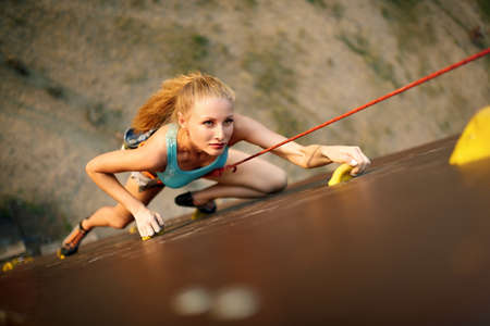 Strong woman and business success concept. Young caucasian pretty woman practicing climbing on artificial rock wall outdoors. Slim sporty blonde training speed bouldering session.の写真素材