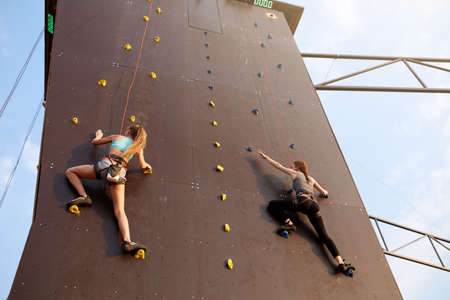 Bottom view of two young climbers approaching to finishing point on the speed track of climbing competitions outdoors. Active sporty women compete on artificial rock wall. Bauldering championship.の写真素材