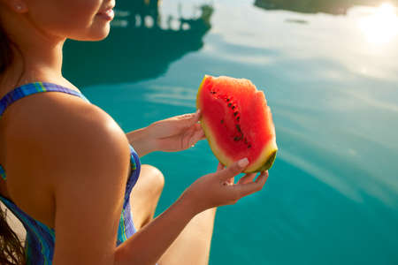 Pretty woman holds slice red watermelon stretching long tanned legs over blue pool, relaxing on tropical island in resort, eating healthy food. Fruit summer diet. Vegetarianism and vegan lifestyle.の写真素材
