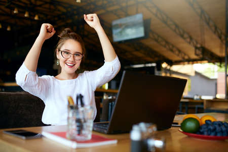 Portrait of happy young business woman celebrating success with arms up in front of laptop. Mixed...
