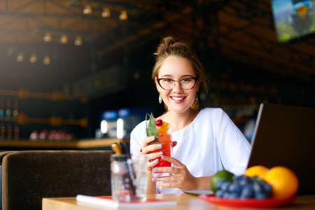 Smiling mixed race woman with cocktail in hand works with laptop. Businesswoman in glasses drinks juice for body hydration while working. Attractive designer quench thirst. Healthy lifestyle theme.の写真素材