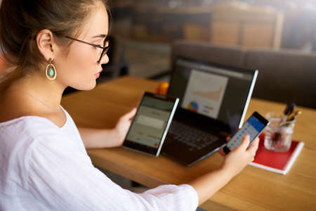 Mixed race woman in glasses working with multiple electronic internet devices. Freelancer...