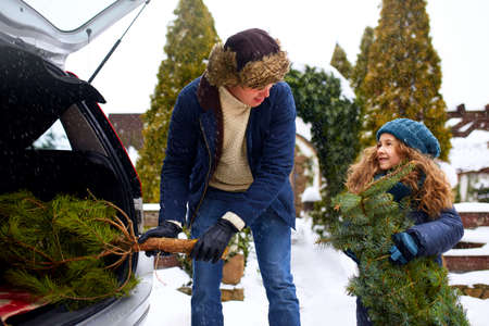 Smiling little girl and father get christmas tree out of car trunk near their house outdoors....