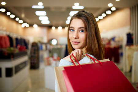 Attractive smiling blonde on shopping in mall. Pretty caucasian girl holding colorfull shopping bags with copyspace in her hand standing in store. Shop lights on background.の写真素材