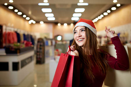 Happy girl shopping gifts in mall on christmas sale. New year holidays shopping idea concept. Smiling pretty caucasian woman with colorful paper presents bags wearing christmas hat in store or shop.の写真素材