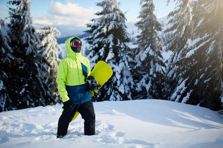 Snowboarder climbing to mountain top carrying his snowboard through forest for backcountry freeride and wearing reflective goggles, colorful fashion outfit and balaclava at ski resort. Winter sports.の写真素材