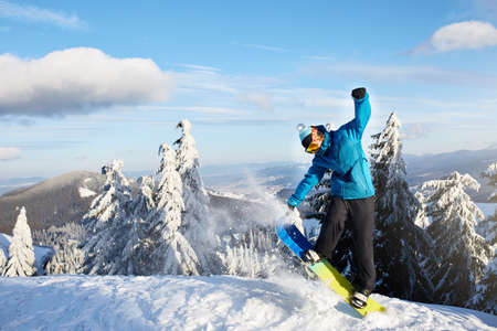 Snowboarder doing tricks at ski resort. Rider performing jump with his snowboard near forest on backcountry freeride or freestyle session in colorful fashionable outfit. Copyspace area. Powder in air.の写真素材