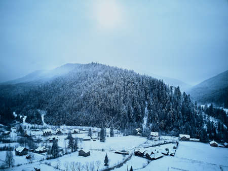 Aerial view of foggy countryside and houses in snowy valley. Hills and mountains with pine tree forest covered in snow. Multi-layered mountain landscape. Ski resort and traveltheme. Moody weather.の写真素材