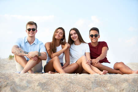 Smiling multiracial friends sitting together on towel on white sand beach during their vacation in an idyllic nature scene destination. Men and women relax on the shore near the sea.の写真素材