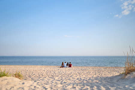 Back view of group of friends sitting together on towel on white sand beach during their vacation and enjoying a sunset above the sea. Men and women relax on the shore with guitar. Tranquil scene.の写真素材