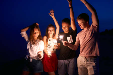 Friends walking, dancing and having fun during night party at the seaside with bengal sparkler lights in their hands. Young teenagers partying on the beach with fireworks. Slow motion steadycam shot.の写真素材