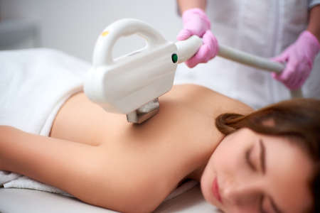 Young pretty smiling woman back laser epilation in beauty salon. Cosmetologist doing depilation treatment with elos device. Beautician hands in gloves holding hair removal cosmetology ipl hardwareの写真素材