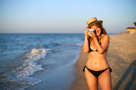Young authentic woman with vintage retro film camera enjoying tropical beach on summer vacation. Female travel photographer in straw hat taking photos having fun at sea. Real girl unretouched shape.の写真素材