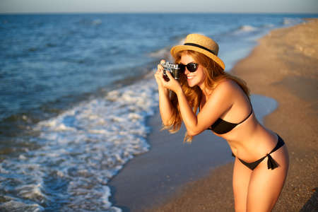 Young authentic woman with vintage retro film camera enjoying tropical beach on summer vacation. Female travel photographer in straw hat taking photos having fun at sea. Real girl unretouched shape.の写真素材