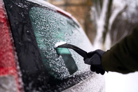 Cleaning the rear car window of snow with ice scraper before the trip. Man removes ice from car rear...