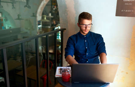 Young attractive hipster freelancer working remotely on laptop in loft cafe. Businessman browsing internet at coffeshop. Blogger posts to social media on travel using 5g internet. Telecommuting theme.の写真素材