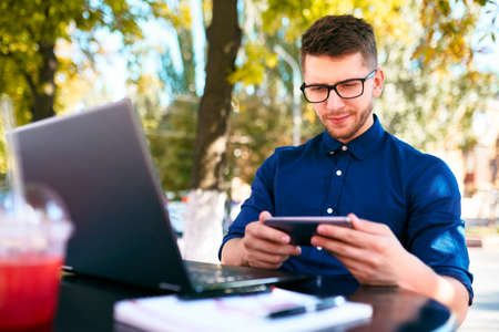 Handsome businessman distracted from work on the laptop watching video on smartphone. Freelancer...