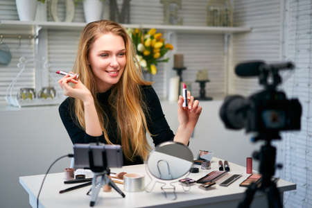 Beauty blogger woman filming daily make-up routine tutorial near camera on tripod. Influencer blonde girl live streaming cosmetics product review in home studio. Vlogger female comparing lipsticks.の写真素材