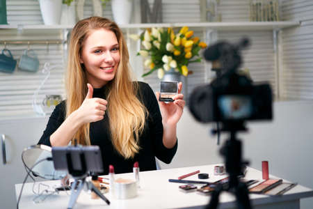 Beauty blogger woman filming daily make-up routine tutorial near camera. Influencer girl live streaming cosmetics product review. Vlogger female recommends eye shadow palette showing thump up sign.の写真素材