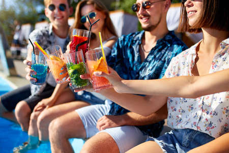Close view. Friends clinking glasses with fresh colorful cocktails sitting by swimming pool on sunny summer day. People toast drinking beverages at luxury villa poolside party on tropical vacation.の写真素材