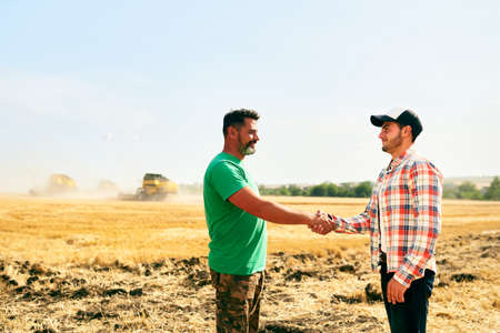 Farmer and agronomist shaking hands in wheat field after agreement. Agriculture business contract concept. Corporate farmer and landlord rancher negotiate with handshake. Combine harvesters harvest.の写真素材
