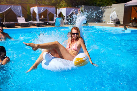 Young smiling bikini girl having fun with friends on inflatable swan in swimming pool. Happy people...