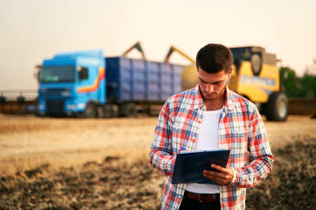 Farmer controls loading wheat from harvester to grain truck. Driver holding clipboard, keeping...