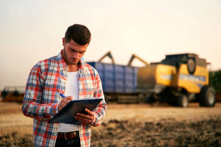 Farmer controls loading wheat from harvester to grain truck. Driver holding clipboard, keeping notes, cargo counting. Forwarder fills in consignment waybills. Agricultural commodities logistics.の写真素材