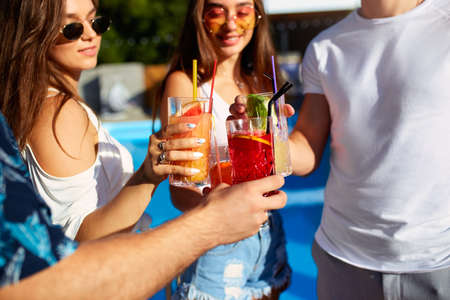 Close view of friends having fun at poolside summer party, clinking glasses with fresh summer cocktails near hotel swimming pool outdoors. People toast drinking juice at luxury villa on vacation.の写真素材
