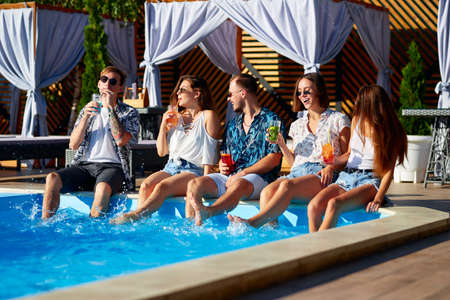 Group of friends having fun at poolside party clinking glasses with fresh cocktails sitting by swimming pool on sunny summer day. People toast drinking beverages at luxury villa on tropical vacation.の写真素材