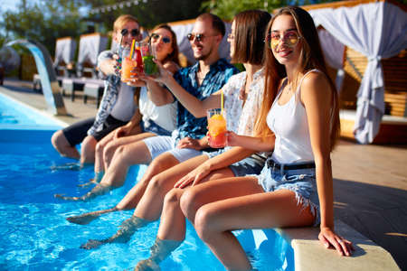 Group of friends having fun at poolside party clinking glasses with fresh cocktails sitting by swimming pool on sunny summer day. People toast drinking beverages at luxury villa on tropical vacation.の写真素材