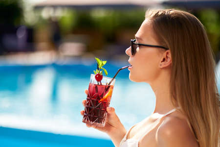 Blonde woman drinks refreshing cocktail sunbathing and sitting near swimming pool at tropical spa....