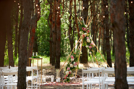 Beautiful bohemian tipi arch decoration on outdoor wedding ceremony venue in pine forest with cones. Chairs, floristic flower compositions of roses, carpet, string fairy lights. Summer rural wedding.の写真素材