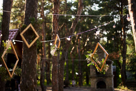 Wedding party banquet outdoors in pine forest. Dining tables, benches decorated in boho style with candles, flowers, spring fairy lights, plates, wineglasses, wooden frames. Eco organic floral decor.の写真素材