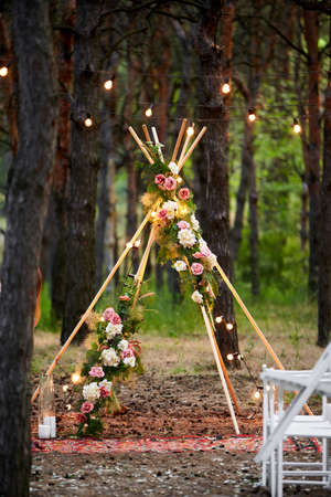 Bohemian tipi arch made of wooden rods decorated with pink roses, candles on carpet, pampass grass, wrapped in fairy lights on outdoor wedding ceremony venue in pine forest. Floristic compositions.の写真素材